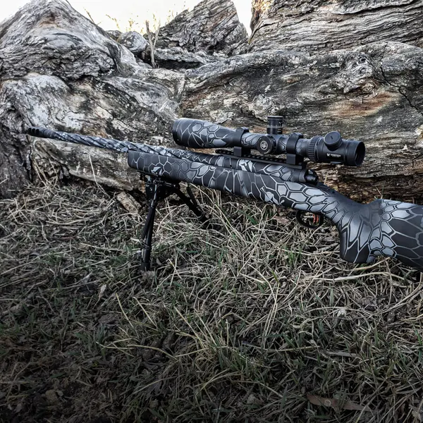 Bolt-action rifle with a black and gray scale-pattern Cerakote camo resting against a log.