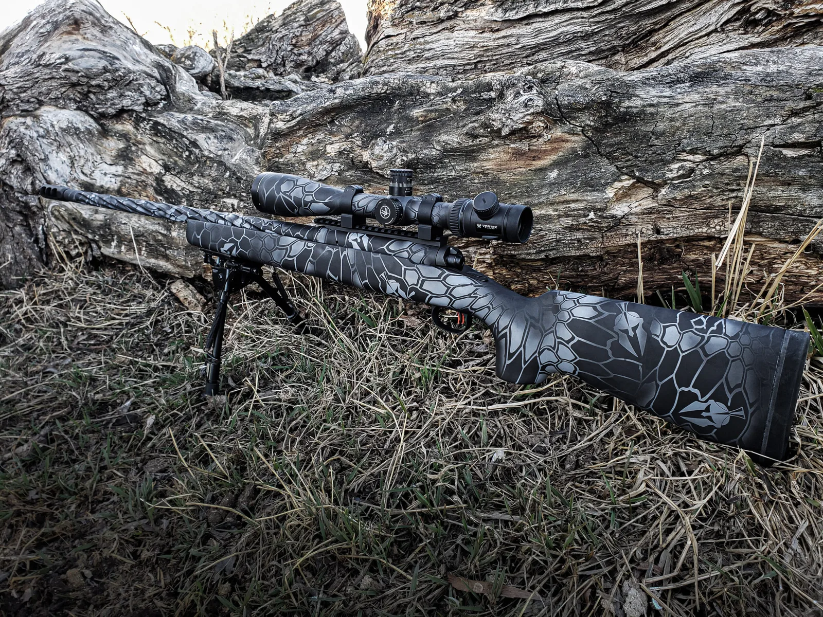 Bolt-action rifle with a black and gray scale-pattern Cerakote camo resting against a log.