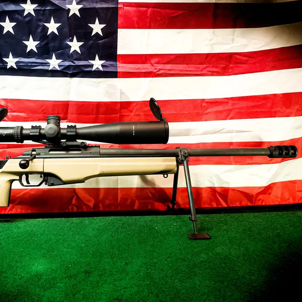 Bolt-action precision rifle with a flat dark earth Cerakote stock and black barrel in front of an American flag.